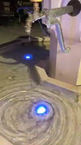 The video shows a water fountain at night. The fountain is made of a light colored stone, possibly concrete. A metal water tap is mounted to the side. The fountain has a pool of water with ripples and two blue lights shining from the bottom. People can be seen blurred in the background.