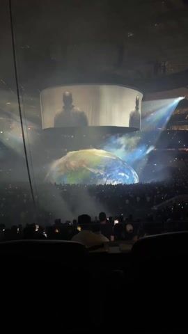 Here is a description of the video:

The video shows a performance, likely a concert, with a large globe-shaped stage in the center.  A figure, presumably a performer, stands on the globe, which has green and blue coloring, possibly representing earth.  Smoke or fog effects are present around the globe.  Above the stage is a large screen showing two figures silhouetted. The audience, holding up cell phones, fills the surrounding stands. There are lights shining on the stage. The lighting is somewhat dim, with bright beams of light shining from various points.