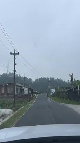 The video shows a point-of-view shot from inside a vehicle, moving forward on a paved road. The sky is overcast and hazy. On the left side of the road are some low-rise buildings and vegetation. On the right side, there are more buildings, some with corrugated metal roofs, and dense green foliage, likely trees on a hillside in the background. Several utility poles with numerous power lines crisscross the sky overhead. A few people are visible along the road; one person appears to be walking or cycling in the distance, and another person is walking on the right side of the road. The overall environment suggests a rural or semi-rural setting, possibly in a mountainous or hilly region.