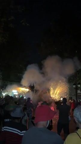 The video captures a night-time outdoor event where people are gathered to watch a display of fireworks. In the center of the frame, a person in a costume, possibly a horse or a creature, is engulfed in a cloud of smoke and sparks from pyrotechnics. The fireworks are erupting around this central figure, creating a dazzling effect of bright light and fire. Several individuals in the foreground and midground are observing the spectacle, some with their phones raised to record the event. The environment appears to be a public space, possibly a street or a square, with trees visible in the background and a dark night sky above. The lighting is primarily from the fireworks themselves, casting a warm, orange glow on the surroundings. The crowd is diverse, with people of various ages and attire, suggesting a community celebration or festival. The overall atmosphere is one of excitement and visual intensity due to the fireworks.