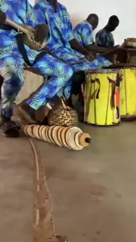 The video shows four African men sitting and playing instruments. They are all wearing the same patterned blue shirts and pants. The man on the left is playing a stringed instrument that looks like a small guitar. The second man from the left is watching the musician. The third man is playing a yellow drum. The fourth man is playing a different drum that is mostly out of view. In front of the men on the floor is a long instrument made of wood and what looks like slices of dried fruit or nuts. A gourd and a stick are also on the floor. The men are sitting on a bench against a white wall in what appears to be an indoor performance space.