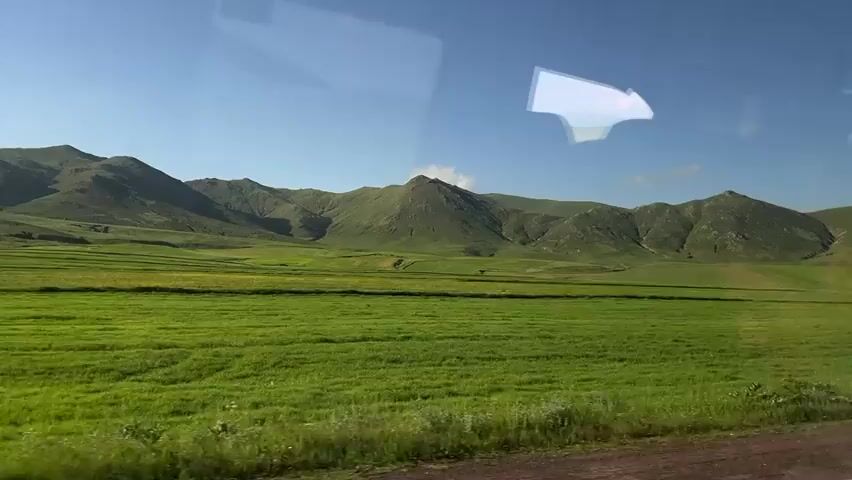 The video shows a view from a moving vehicle, looking out at a green, rolling landscape. The foreground consists of a green field, with a darker road or dirt path at the very bottom of the frame. In the middle ground, the green field continues, and further back, there are rolling hills and mountains, some with patches of green and others appearing more barren. The sky is blue with a few wispy clouds. There is a reflection on the vehicle's window, showing a white, arrow-like shape and a distorted image of the sky and hills. There are a few yellow wildflowers visible in the foreground. A white road marker is visible to the right in the middle ground. The overall impression is of a rural, natural landscape.