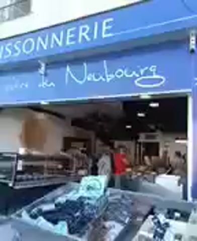 The video shows the exterior of a fish market. The market has a blue awning with "POISSONNERIE" and "Le Neubourg" written on it in white cursive letters. There is also a logo of a crab on the awning. In front of the market, there are displays of fish on ice. Two people, a man in a red shirt and a person in a light-colored shirt, are visible inside the market. The video is shaky and out of focus.