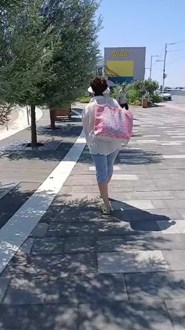 The video shows a person walking away from the camera on a paved walkway. The person is wearing a white shirt, light blue shorts, and flip-flops. They are carrying a pink and white striped bag over their left shoulder. The walkway is lined with trees and has a white stripe running along its edge. In the background, there is a building with a yellow sign that says "PIRAEUS". Another person is walking in the opposite direction in the distance. The sky is clear and blue, indicating it is a sunny day. The lighting suggests midday.