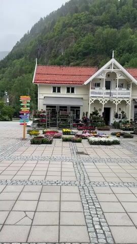 A daytime video shows a charming building with a light yellow facade, red tiled roof, and white decorative trim, situated against a backdrop of a lush green, misty mountain. The building appears to be a flower shop or bistro, with numerous pots and trays of colorful flowers (red, yellow, white, pink, purple) displayed outside on a paved courtyard. The courtyard features a pattern of square light-colored pavers and darker cobblestones.

To the left, a colorful signpost with directional arrows stands. To the right, a black A-frame sign reads "bistro ALEN" with a logo, "OPEN BISTRO," "FOOD," "DRINKS," "ICE CREAM," and "WELCOME." A white canopy tent and a red bicycle with a flower basket are also visible near the bistro sign. A woman in a white sweatshirt walks from the left side of the frame towards the right, crossing the paved area. The camera pans slightly to follow her movement. The scene suggests a quaint, welcoming atmosphere, likely in Dalen, Norway, as indicated by the sign.