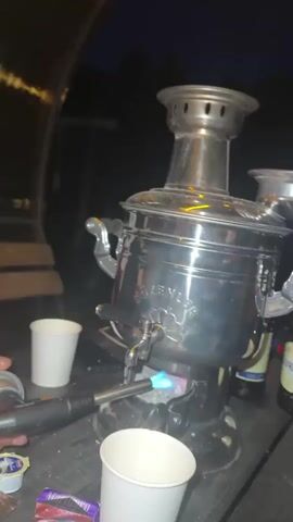 The video shows a close-up, slightly shaky shot of a metallic samovar on a wooden table at night. A torch is being used to ignite the fire beneath the samovar, producing a blue flame. There are several white paper cups on the table, along with a pack of cigarettes and some small packets that appear to be creamer or sugar. In the background, the dark shapes of trees are visible, suggesting an outdoor setting, possibly a campsite or backyard. The lighting is dim, with the flame from the torch and the reflection on the samovar being the primary light sources. A portion of a person's arm, holding the torch, is visible in the lower left corner of the frame.