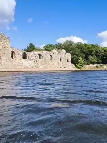 The video shows a wide body of dark blue water with choppy waves. In the background, there is a large stone ruin of a castle or ancient building. The ruin is made of rough-hewn stones and has several arched openings. The top of the ruin is crumbling. Behind the ruin, there are lush green trees and a blue sky with white, puffy clouds. The camera appears to be moving slowly, possibly from a boat.