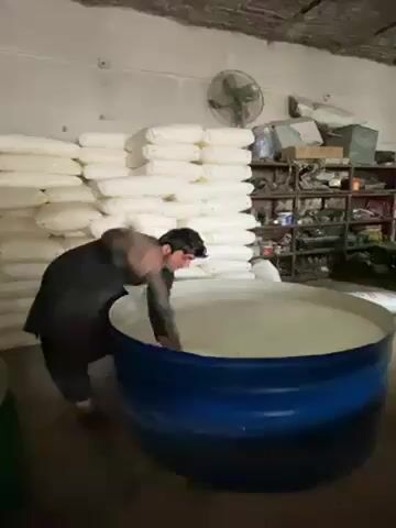 A man is mixing the contents of a large blue barrel using a wooden paddle. The barrel is full of a white substance. He is wearing a long-sleeved black shirt. Behind him, there is a stack of white bags. To the right, there is shelving with various items. A fan is mounted on the wall in the background. The room appears to be a warehouse or industrial space.