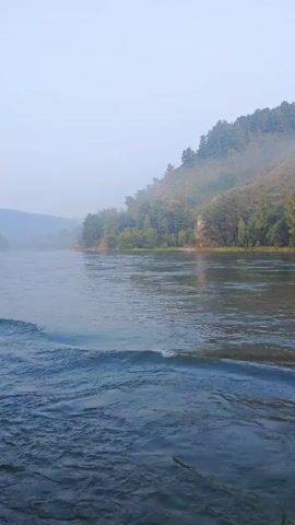 A calm river flows past a steep, rocky cliff face covered in vegetation. A light mist hangs in the air, obscuring the distant hills. The water is dark and reflective, with small waves breaking on the surface.
