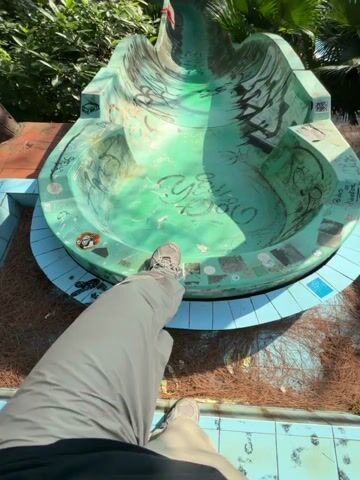 A first person point of view shot shows a person standing on an old green and blue painted water slide, then walking up the slide. The slide is covered with graffiti. The slide curves into a tunnel covered in spray painted murals. There are trees and vegetation surrounding the slide.