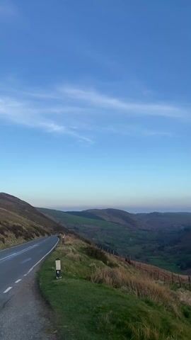 The video shows a wide-angle view of a landscape featuring rolling hills and a valley, captured under a clear blue sky with sparse clouds.  A road is visible on the left side of the frame, curving along the hillside.  The valley below is lush and green, with hints of rural development visible in the distance.  In the foreground, a grassy area is delineated by a simple wooden fence. The hills in the background are covered in a mix of vegetation, appearing brown and green.