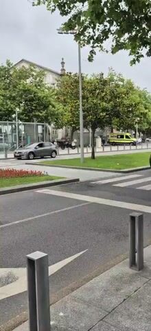 The video shows a grey car driving past a bus stop with a glass shelter. In the background, there are trees and a building. The video then pans to show a red van and more trees. Next, a black car is parked in front of a building with a dome. Finally, the video shows a white electric car driving on a roundabout, followed by a black car driving past a building with columns.