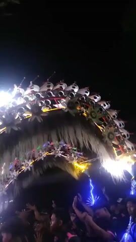The media file depicts a nighttime scene at what appears to be an outdoor event or festival in Dukuh Klopo, Indonesia. The primary focus is on a large, elaborate archway adorned with white, fluffy material, resembling hair or fur, and decorated with numerous colorful lights, including blue, green, pink, and yellow. The archway is further embellished with star-shaped ornaments and what seem to be circular, metallic objects arranged in a pattern. Hanging from the top of the arch are what look like small, light-up figures or ornaments.

Beneath and around the archway, a crowd of people is visible, many of them wearing dark clothing, some with pink accents. They are standing, watching the spectacle, and some have their hands raised in the air, suggesting they are engaged and enjoying the event. Bright lights, including spotlights and string lights, illuminate the scene, creating a festive and energetic atmosphere. The background is dark, indicating it is nighttime, and there are no obvious weather cues.

The overall activity appears to be a procession or a ceremony, possibly a parade, given the scale of the decorated archway and the gathering of people. The bright lights and the decorated structure suggest a celebratory occasion. There is no discernible text visible in the media file. The emotions conveyed are those of excitement and festivity, common in local celebrations.