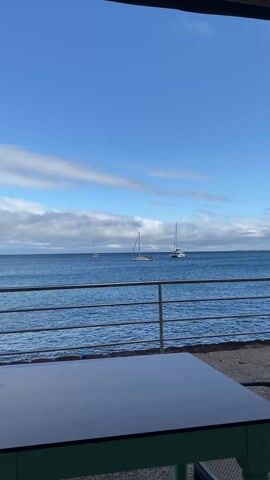 The video depicts a view of the ocean from what appears to be a restaurant or seaside establishment. In the distance, three sailboats are visible on the water under a blue sky with scattered white clouds. The foreground features a metal railing, a table with a dark top, and a green base. The shot is framed by the doorway or window of the structure, partially obscuring the top and sides of the view. The ocean appears calm, with gentle waves. The light suggests it is daytime. Sounds of people talking are audible.