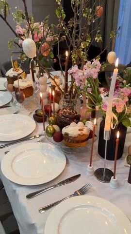 The video shows an Easter dinner table. The table is set with white plates, silverware, candles, Easter eggs, and flowers. There are multiple cakes on the table. A person is seen in the background.