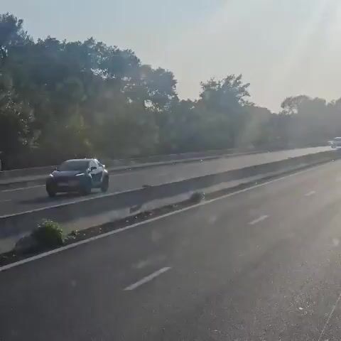 The video shows a highway with multiple lanes of traffic. The sun is shining, creating a bright, hazy atmosphere. There are trees and foliage lining the side of the highway. Several vehicles are visible, including cars and trucks. One truck appears to be a fire truck, distinguished by its red color and emergency lights. The vehicles are moving along the highway, suggesting normal traffic flow. The camera appears to be mounted on a moving vehicle, providing a first-person perspective of the scene.