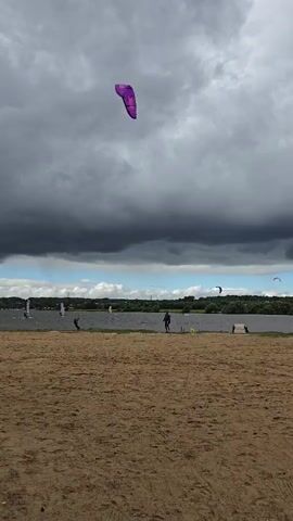 A wide shot shows a beach with sand in the foreground. In the middle ground, a body of water is visible, with a line of trees on the opposite shore. Several windsurfers are on the water, and a person stands on the beach near the water. In the sky, a purple kite is visible. The sky is filled with dark, dramatic clouds.