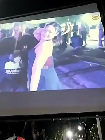 A projection screen displays a woman in a strapless dress dancing. The screen is illuminated by bright lights, creating a blue and white effect. In the background, blurred figures and what appears to be a festival or outdoor event are visible. The lower portion of the frame shows a dark area, possibly the interior of a venue or the audience watching the projection. The video is shaky and blurry.