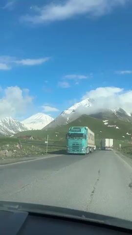 A turquoise Volvo truck is seen on a road with snow-capped mountains in the background. Other vehicles, including white SUVs and other trucks, are also present on the road. The sky is blue with white clouds. The video appears to be shot from inside another vehicle.
