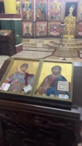 A vertical video shows the interior of a brightly lit church or cathedral. In the foreground, two framed religious icons featuring figures with halos are displayed on a wooden surface. Small bills of money are placed on the icons. The camera then pans up and to the left, revealing more of the church's ornate interior. The walls are adorned with numerous religious paintings and frescoes, many depicting saints and biblical scenes. A large, golden chandelier with multiple lit candles hangs prominently from the ceiling, which is also painted with religious imagery. The floor has a decorative tile pattern. The video ends looking up at the chandelier and painted ceiling.