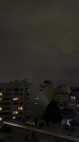 The video depicts a nighttime cityscape under a heavily clouded sky. Several multistory buildings are visible, their windows illuminated with warm-toned lights. A dark, leafy tree stands in the foreground amidst the buildings. The overall scene is dimly lit. No people or actions are directly observable.
