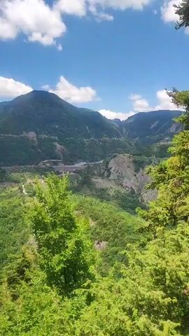 This is a vertical video captured from a high vantage point, overlooking a lush, mountainous landscape. The foreground is dominated by vibrant green trees, with a tall, slender evergreen tree on the left and a denser cluster of foliage on the right.

Beyond the trees, the scene opens up to a vast valley. A winding road or river is visible snaking through the valley floor, surrounded by more green, densely forested hills. In the distance, larger, more rugged mountains rise up, their slopes partially obscured by shadow and patches of lighter rock or earth. The scale of the mountains suggests a significant elevation.

The sky above is a brilliant blue, dotted with fluffy white clouds. The lighting indicates it is daytime, likely mid-morning or afternoon, with bright sunshine illuminating the scene. The overall impression is one of natural beauty, tranquility, and a sense of vastness. There are no people or man-made objects prominently featured, emphasizing the wild and scenic nature of the location. The setting appears to be a rural, mountainous region, possibly a national park or a protected natural area, given the pristine condition of the greenery. The visual elements evoke a feeling of peace and awe at the grandeur of nature.