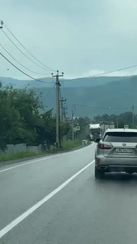 The video shows a view from inside a car driving on a wet road in a mountainous, rural area. Overhead power lines are visible, running along the side of the road. There are several utility poles with multiple wires. Trees and vegetation line the left side of the road, and a metal fence is also present. On the right side of the road, a gray Lexus SUV is driving in front of the camera car. Further ahead on the road, there are pedestrian crossing signs and a large white semi-truck is approaching. The sky is overcast, and the overall environment appears damp, suggesting it may be raining or has recently rained. The mountains in the background are covered in greenery and mist.