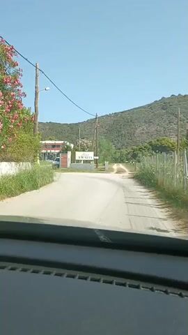 The video shows a view from inside a vehicle, looking out at a sign that reads "Hotel AMALIA". The sign is white with blue lettering and is flanked by grey pillars. A dirt road leads away from the sign into a grassy area, with a green fence on the right. In the background, there are trees and a hill covered in vegetation under a clear blue sky. To the left of the sign, there is a building with a red roof. The vehicle appears to be approaching the hotel entrance.