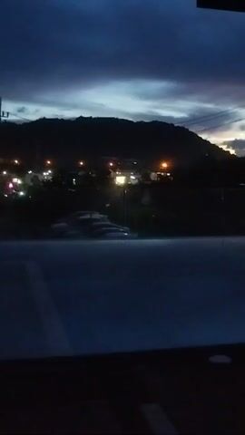 The video shows a dark, outdoor scene at dusk. In the distance, a mountain range is silhouetted against a twilight sky, with scattered lights indicating a small town or settlement. A telephone pole with wires is visible on the right side of the frame. In the foreground, there is a dark, reflective surface, possibly a body of water or a polished floor, which mirrors the dim lights and sky. A large, dark pillar stands vertically in the middle right of the frame, partially obscuring the view of the landscape. The overall impression is one of a quiet, possibly serene, evening setting viewed from a sheltered vantage point.