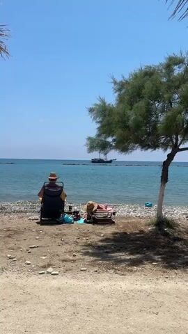 A medium shot depicts two people enjoying a sunny day at the beach. One person, wearing a hat, is seated in a black chair facing the ocean. Nearby, another person reclines on a beach chair. Several items are placed on the pebbly sand between the chairs. In the distance, a large sailing vessel is visible on the water. The beach is sandy with some pebbles. A tree stands to the right of the chairs, casting a shadow on the ground. The sky is clear and blue. The camera is static.