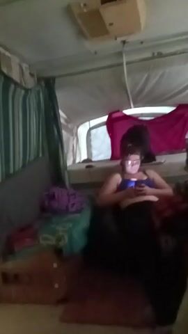 The interior of a pop-up camper is shown. A young woman with glasses is reclining in the bed, looking at her phone. A pink shirt hangs over the back of the bed. There is an air conditioning unit mounted on the ceiling. Striped curtains are on the left side of the image. The woman speaks after being asked a question.