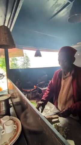 An elderly man, wearing a red cap and a red jacket over a light-colored tunic, is preparing food in a small, open-air food stall. He stands behind a counter, in what appears to be a mountain setting, with a window behind him offering a view of lush greenery and a tall conifer tree. The sky is overcast, suggesting it might be morning or a cloudy day. The man is actively engaged in cooking; a pan on a stove in front of him is alight with flames, and he is using a rolling pin to flatten something on a wooden surface, with chopped ingredients and a block of white cheese nearby. To his left, there are a few white teacups stacked on a decorative plate and a stainless steel container. Two woven lampshades hang from the ceiling of the stall, casting a warm glow. A glimpse of a sign with yellow lettering is visible through the window, but the text is indistinct. The overall impression is one of a humble, authentic culinary experience in a scenic, possibly rural, location.