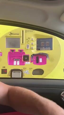 The video begins with a close-up of a person's hand inserting a red credit card into a yellow payment machine. The machine has a screen displaying "Veuillez patienter s.v.p.", indicating "Please wait". Below the screen, it shows "Classe 1" and "Prix : 8.00 €", meaning "Class 1" and "Price: €8.00". To the left, there are slots for coins and bills, and a pink area for receipts. The view then shifts to the interior of a car, showing the payment machine through the driver's side window and a side mirror reflecting the hand and card. Finally, the video shows the view from inside the car looking forward, as a red car drives away from a toll booth under a cloudy sky.