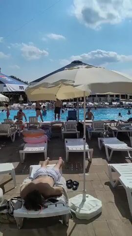 The video shows a sunny day at an outdoor pool party. In the foreground, several empty white lounge chairs are arranged on a paved area. One lounge chair in the foreground has a person lying face down on it, wearing a white patterned shirt and shorts. Another lounge chair has a person's black flip-flops on it. In the middle ground, people are seated on lounge chairs and standing around, some are under umbrellas. A large Pepsi branded umbrella is visible. The background shows a pool area with many people gathered, and a foam machine is producing a large cloud of white foam in the air, creating a festive atmosphere. People are interacting in the foam, some dancing and some observing from the poolside. The sky is blue with scattered white clouds.