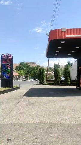 The video shows a gas station in a sunny, outdoor environment with a clear blue sky and some scattered clouds. The camera pans slightly to the right, revealing a digital price display board for LPG, with prices listed as 240, 240, and 550. Further to the right, a red and white awning of the gas station structure is visible. Across the street, there are buildings, trees, and several cars, including a silver sedan in motion. The ground is paved, likely asphalt. The overall scene suggests a typical roadside gas station in a town or rural area.