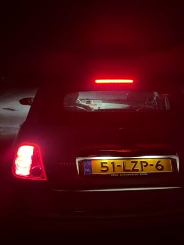 The video begins with a close-up of the rear of a dark-colored car at night. The license plate, which reads "51-LZP-6" with a Dutch flag emblem, is clearly visible. The car's brake lights and taillights are illuminated, casting a red glow. A faint reflection of a person can be seen in the rear window. The camera then pans to the left, revealing the car's headlights cutting through the darkness. The scene transitions to a wide shot of a dark forest silhouetted against a night sky. A bright full moon is visible above the trees, partially obscured by scattered clouds. The overall atmosphere is dark and quiet.