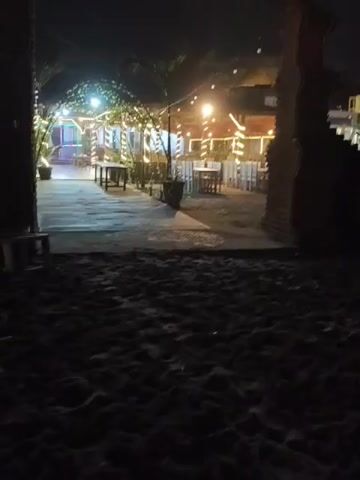 The video shows a dimly lit outdoor area at night. The foreground is dark sand, presumably a beach. Beyond the sand, there is a paved pathway leading towards a seating area with tables and chairs. The area is decorated with numerous string lights, creating a festive atmosphere. The seating area appears to be an open-air restaurant or cafe, with thatched roofs visible in the background. There are no people visible in the video. The lighting from the string lights casts a warm glow on the scene, and some palm trees are visible in the background, suggesting a tropical location. The overall impression is one of a tranquil, inviting nighttime venue.