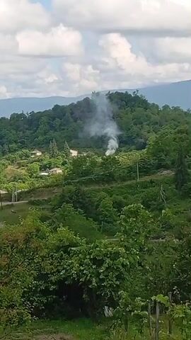 A smoke plume rises from a wooded hillside, obscuring houses nestled among the trees. The smoke appears to be coming from a small fire or smoldering material. The surrounding landscape is characterized by lush green vegetation, rolling hills, and a cloudy sky. The scene suggests a rural or semi-rural setting.