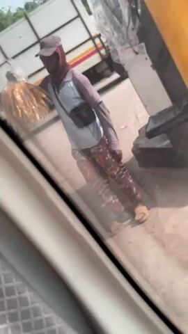 The video shows a person seen through a car window. The person is wearing a grey cap, a brown knitted face covering, a light blue short-sleeved shirt over a grey long-sleeved shirt, and floral pants. They are also wearing sandals and a black crossbody bag. The person is holding a bag full of what appears to be golden-brown snack food. A white truck with red and yellow stripes is visible in the background. The car window has a white frame and the interior shows a textured grey surface. The angle is slightly tilted, making the person appear to lean to the left.