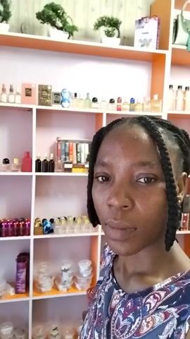 A woman with dark braided hair stands in front of shelves stocked with perfumes and other cosmetic products. She is wearing a patterned top and looking at the camera. The shelves are organized and well-lit, displaying a variety of bottles and containers. The background suggests a retail environment or a personal collection of fragrances. The lighting is bright, casting a soft glow on the scene.