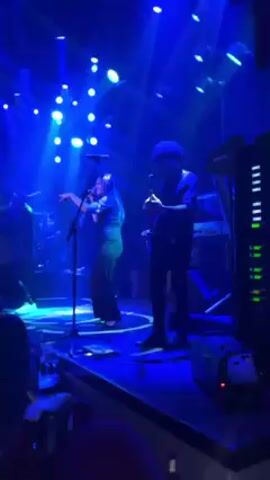 The video shows a band performing on a small stage in a nightclub. The stage is brightly lit with blue stage lights. There are four band members visible on stage: a male singer on the left, a female singer in the center, a guitarist on the right, and another male singer further back. The female singer is wearing a dark top and pants. The guitarist has dark curly hair. There are audience members visible in the foreground, seated at tables in front of the stage. The band is playing enthusiastically, and the audience appears to be enjoying the performance.