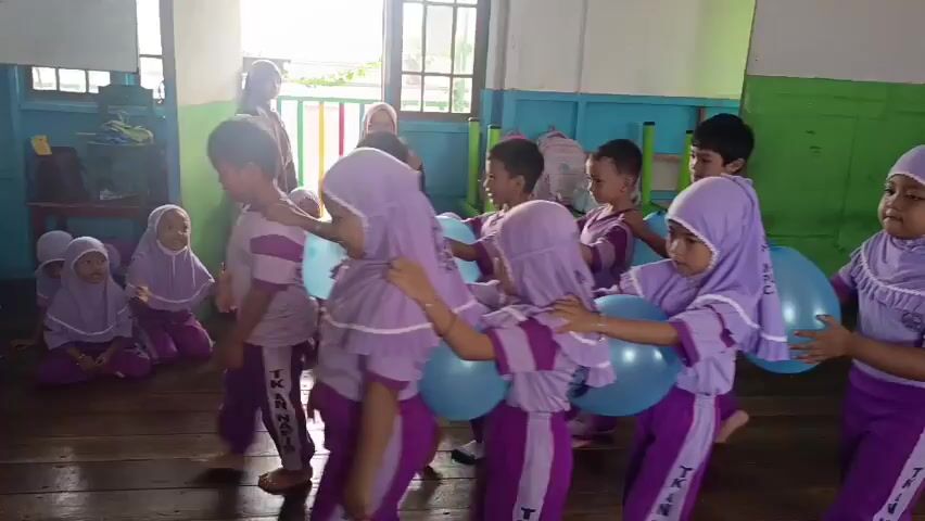 A group of young children, appearing to be in a classroom or community center, are participating in a game involving balloons. The children, dressed in matching purple and white uniforms with headscarves, are arranged in a line and are holding large, light blue balloons between their bodies, specifically between their backs and arms. They are all facing the same direction and seem to be moving together as a unit. Two adult women are also present in the room. One woman, wearing a black hijab and a beige skirt, stands to the left, holding a microphone, and appears to be addressing the children. Another woman, wearing a red top and a black hijab, stands further back in the room, observing the activity. The environment appears to be a simple room with wooden floors and walls painted in various colors, including green and blue. Sunlight streams in from a window on the right side of the frame, illuminating the scene. The overall impression is of a playful and organized activity for young children.