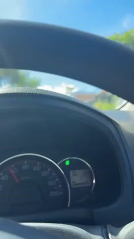 The video shows the view from the driver's seat of a car. The dashboard is visible, with the speedometer reading approximately 20 km/h and the odometer reading 215. A green indicator light is illuminated. The road ahead is paved with a yellow dividing line. On either side of the road are houses and trees, with a clear blue sky and some clouds overhead. A black car is visible in the distance, traveling in the same direction. The sun is shining brightly.