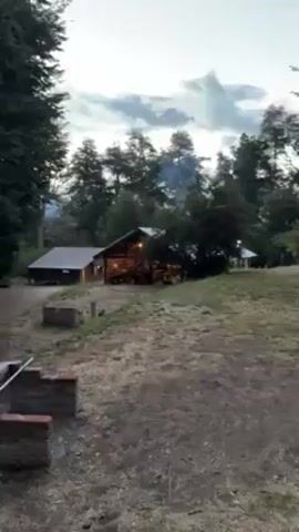 The video depicts an outdoor setting at dusk, with a large wooden structure serving as a focal point. Smoke is visible rising behind it, suggesting a fire. Several individuals are gathered inside or around the structure, although their activities are unclear due to the distance. The surrounding environment includes grassy terrain and tall trees, contributing to a natural, wooded atmosphere. A small brick structure is visible in the foreground. The sky is overcast with some visible sunlight peeking through the clouds.