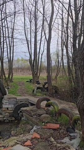 The camera pans over a small stream meandering through a forest. The stream is littered with old tires. The stream is spanned by a makeshift bridge made of cinder blocks and other discarded materials. The trees are mostly bare, suggesting it is either late fall or early spring. The overall atmosphere is one of neglect and environmental degradation.
