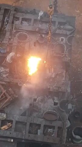 A high-angle, full shot shows a large, flat metal object on the ground. It appears to be a piece of industrial machinery, possibly the hull of a tank, with numerous circular openings and rectangular compartments. A person in protective gear, including a helmet and possibly a welding mask, is operating a torch on the metal object, creating sparks and flames. A chain and hook from an overhead crane are suspended above the object, with the hook attached to one side. There are scattered metal scraps and containers around the object. The overall environment appears to be an industrial setting, possibly a workshop or scrapyard. The lighting is dim, with the primary light source being the intense glow of the torch.