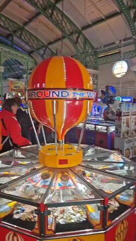 The video depicts the interior of an arcade, characterized by a high ceiling with ornate green ironwork and a variety of arcade games. The games include coin-operated machines and claw machines, with bright neon lights adding to the atmosphere. People are seen moving around the arcade, including individuals wearing casual clothing. There are ticket machines and a change machine for obtaining coins. The camera pans across the scene, showcasing the arcade's layout and diverse range of games.
