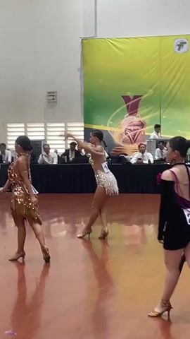 Three young women in sparkly dance costumes are performing a synchronized dance routine on a polished wooden floor in what appears to be a competition. A large banner with a yellow and green design hangs in the background, featuring a trophy graphic and text in an Asian language. A panel of judges sits at a long table in front of the banner. The dancers are wearing short, fitted dresses adorned with sequins and fringe, and high-heeled dance shoes. Their hair is pulled back. The lighting is bright and even, typical of an indoor event space.