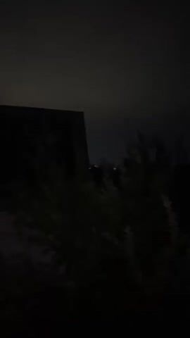 The video shows a dark, nighttime scene with a blurry, bright object in the center, possibly the moon or a light source, partially obscured by foliage. The sky is dark and cloudy, with a faint glow. In the background, there are silhouetted shapes of buildings or structures. The overall impression is a dimly lit, outdoor environment at night.