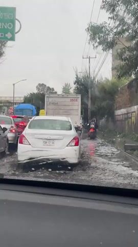 The video shows a traffic jam on a street during rainfall. Several cars are stationary in what appears to be flooded conditions, with water covering the road surface. A white sedan is prominently featured in the foreground, with its rear facing the camera. Behind it, a large white truck is visible, along with other vehicles in the traffic queue. A person on a motorcycle is navigating through the water and traffic on the right side of the road. The environment appears to be urban or suburban, with buildings and trees lining the street. The sky is overcast, indicating continuous rain. The overall scene depicts a challenging driving situation due to the weather and road conditions.